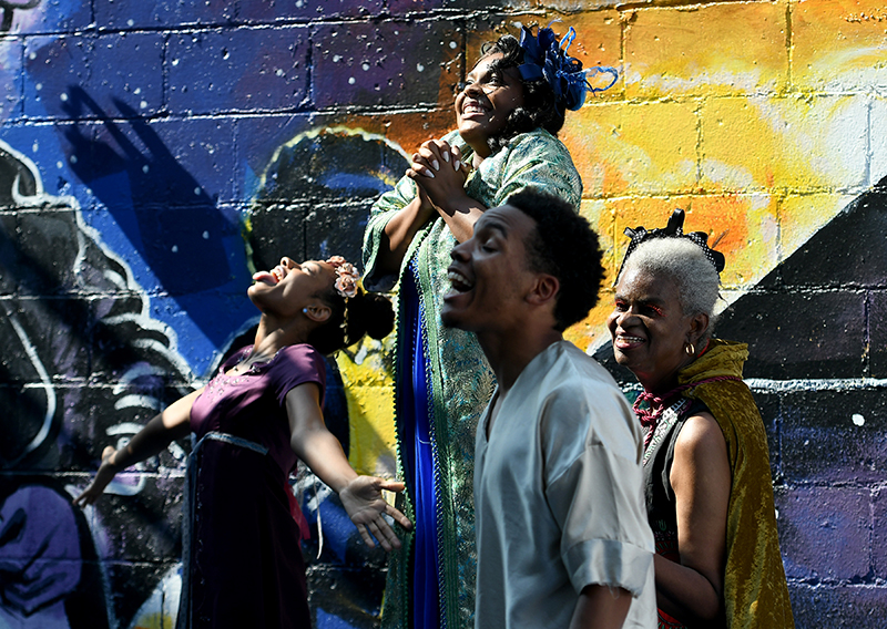 Performers from Black and Brown Theatre perform in front of a wall with a vibrant mural at a sidewalk festival.