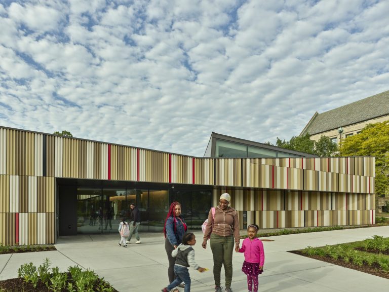 Two adults, presumably parents, holding the hands of a small child each on the walkway leading to the terra cotta clad Marygrove Early Education Center.