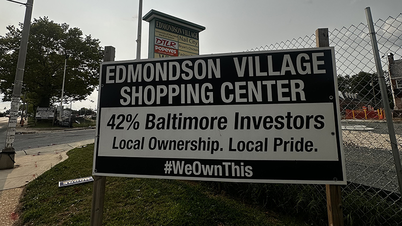 A large sign sits in front of a fence with a shopping center lot behind it, Sign reads: Edmondson Village Shopping Center | 42% Baltimore Investors | Local ownership. Local Pride.