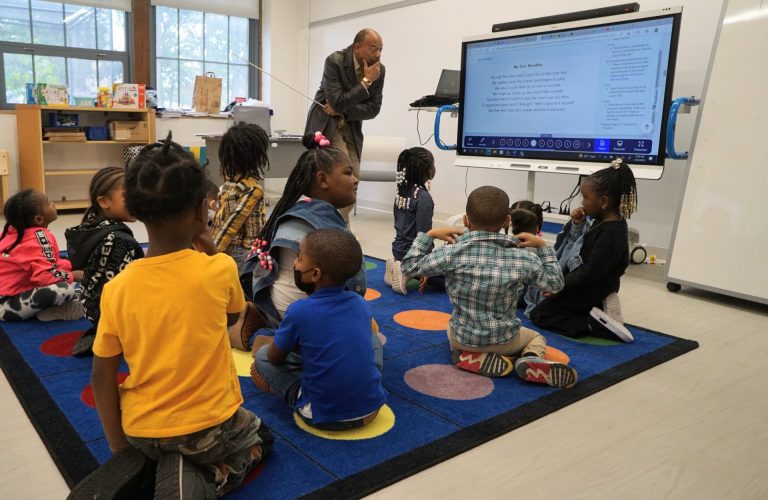 Teacger with yard-long pointer looks attentively to a lesson on a large computer monitor with a classroom of 10 or so young students (perhaps second graders) seated on a rug looking on.