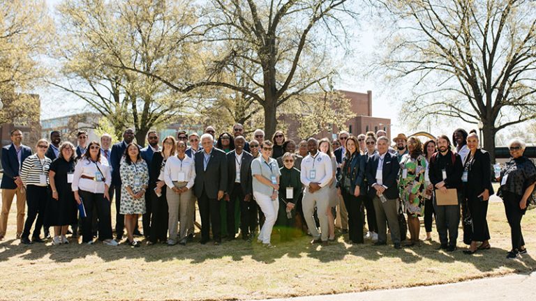 A large group of about 40 people stand together in park in front of several trees as they pose for a photo.