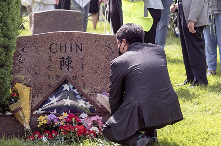 A man in a suit and a mask kneels down in front of the tombstone of Vincent Chin. 