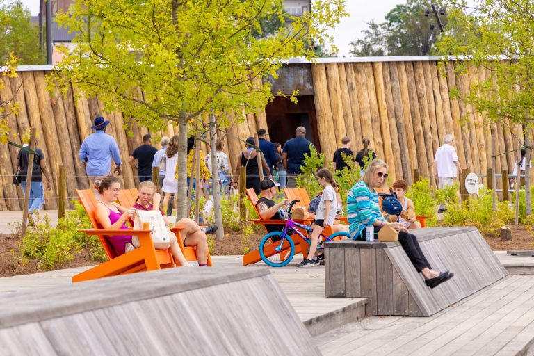 People are seen sitting in adirondack chairs, walking on trails along a bamboo wall