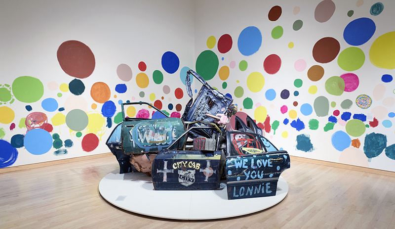 A mixed media installation that features a series of car doors painted with images and slogans seen against a wall full of colorful polka dots.