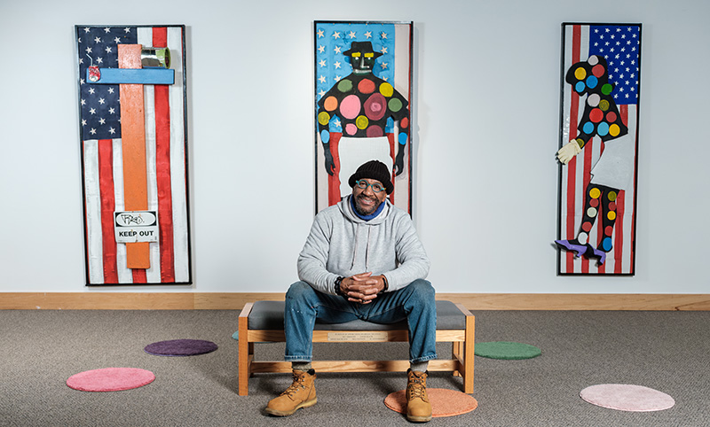 2026 Kresge Eminent Artist Tyree Guyton in a grey sweatshirt and knit cap sits on a bench in front of three of his artworks on the wall behind him that features flag images and polka dots. 