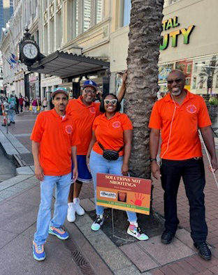 Four people wearing orange Ubuntu Village shirts on a street in New Orleans standing next to a tree and a sign that says Solutions not Shootings. 