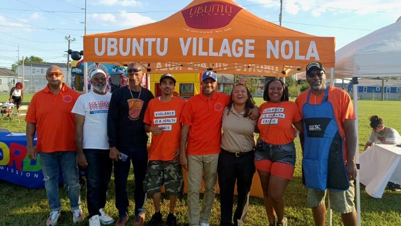 A group of eight people stand together in a field in front of a tent with an orange cover that says Ubuntu Village NOLA. Several of the people are wearing orange Ubuntu Village shirts that say Violence is a health crisis.