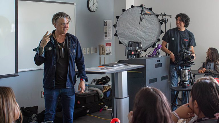Two men are standing in front of a classroom of students with filmmaking equipment around them.