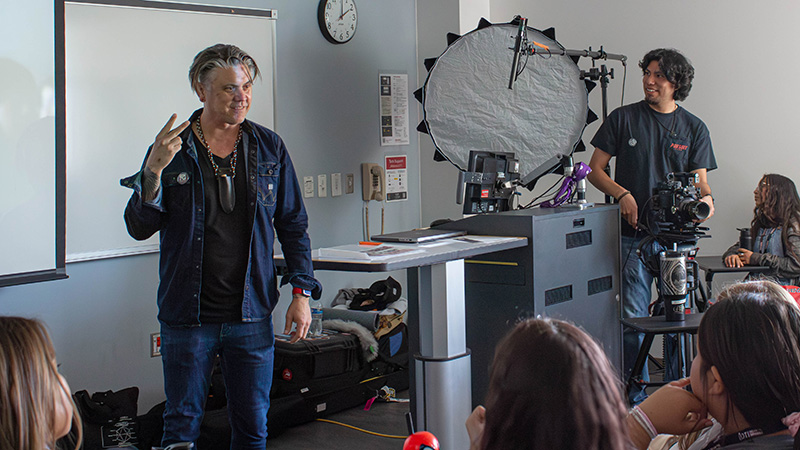 Two men are standing in front of a classroom of students with filmmaking equipment around them.