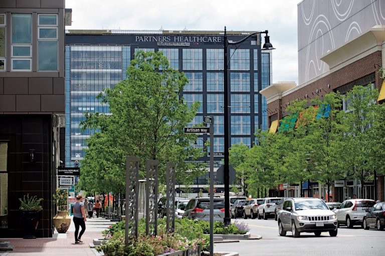 The image shows a city street, with green trees on each side. At the end of the street is a black office building with the words "Partners Healthcare" at the top.
