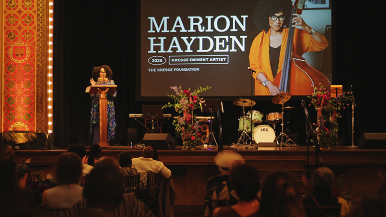 A Black woman stands at a podium on a stage with a small band set-up with instruments and a screen in the back with a image of her playing the bass and the text: Marion Hayden, 2025 Kresge Eminent Artist.