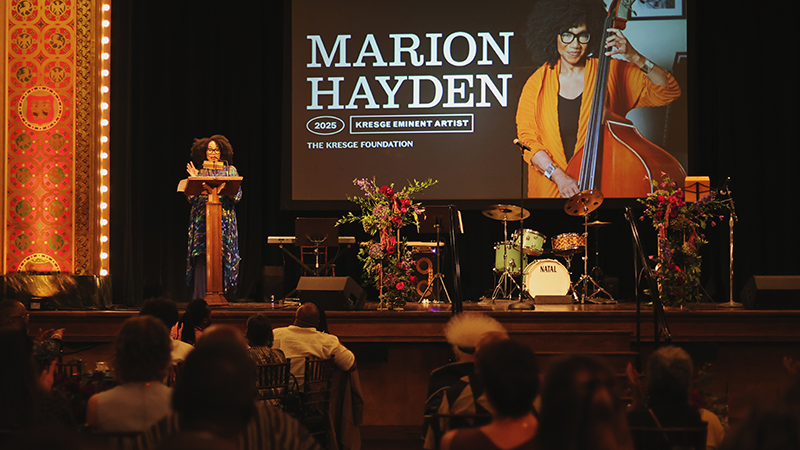 A Black woman stands at a podium on a stage with a small band set-up with instruments and a screen in the back with a image of her playing the bass and the text: Marion Hayden, 2025 Kresge Eminent Artist.