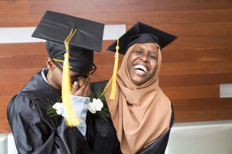 A male wearing a black graduation gown holds his face while being embraced by a woman wearing a brown headscarf and a black graduation gown.