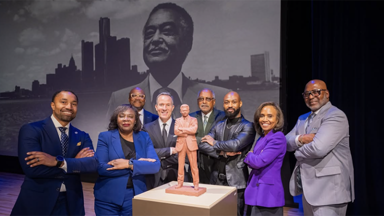 A group of people are gathered around a model of a statue for the proposed Colema A. yung tribute. ALl the people are standing with their arms acrosee their chest in the same fashion as the statue.