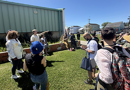 A group of people gathered in a green space next to a building listen to a man talking.