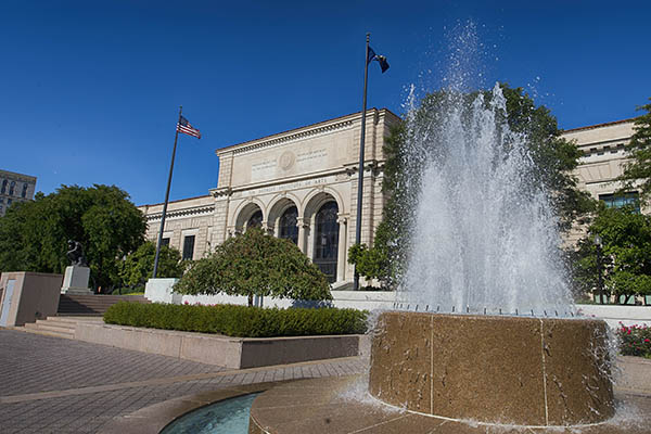 The exterior of the Detroit Institute of Arts, a large museum with a fountain and trees in a plaza in front of it.