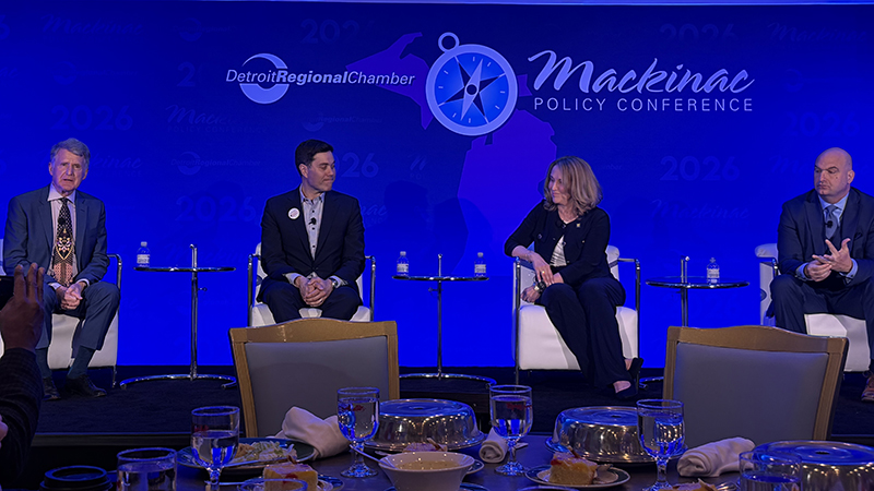 Three men and one woman sit in white chairs on a stage with a blue background with the words: Mackinac Policy Conference.