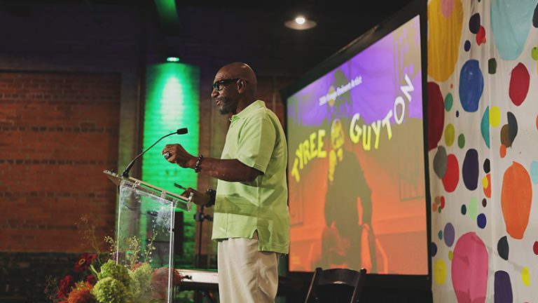 2026 Eminent Artist Tyree Guyton in alight green shirt stands at a podium on a stage with a screen with an image of him and a wall covering with colorful polka dots.