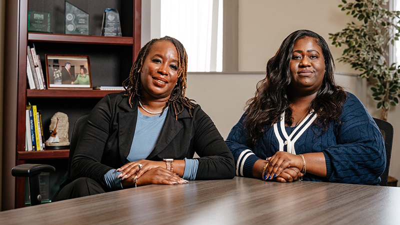 Two Black women are sitting at a table and smiling wutg a bookcase and green plant behind them.
