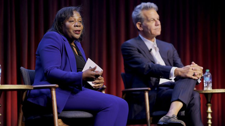 A Black woman and a white man sit in chairs on stage with a red curtain behind them as part of a panel at a conference