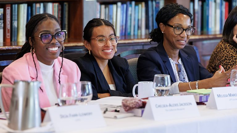 Three women of color are seated behind a table with their names displayed in front of them.