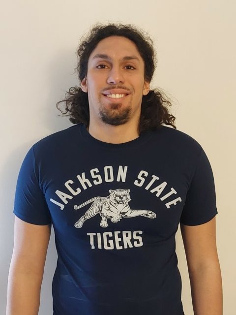 A light-skinned Black man with shoulder-length hair that is half pinned back smiles and wears a Jackson State Tigers tee shirt.