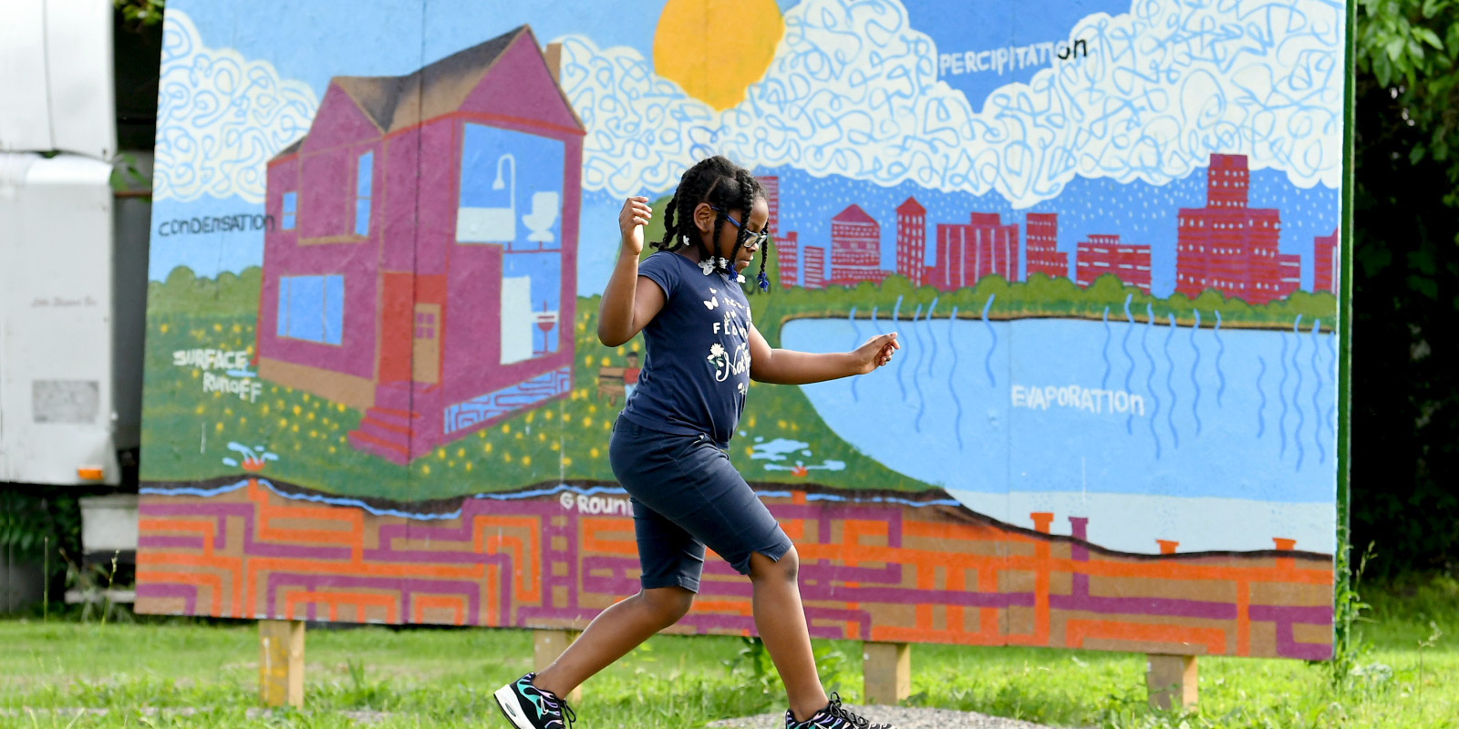 A young girl skips over a tree trunk with a mural of a red house next to a pond with buildings in the background.