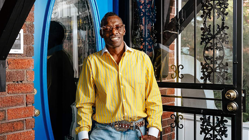 A Black man in a yellow shirt is smiling and standing outside of his house next to a blue front door with a window.