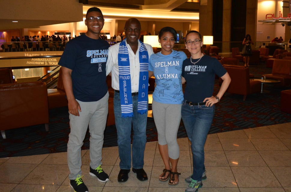Four individuals stand in a reception hall smiling at the camera with their arms around one another, wearing Spelman College shirts.