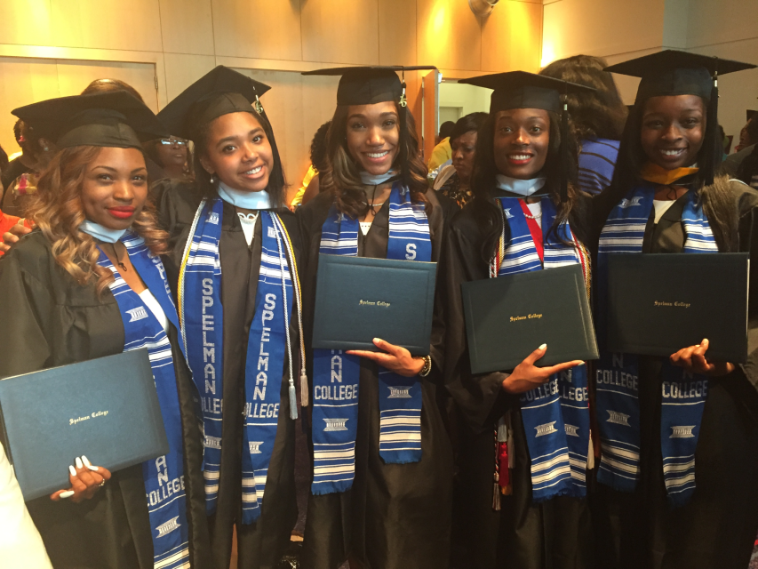 Five Black woman stand shoulder to shoulder in graduation caps and gowns, holding their degrees up for the camera.