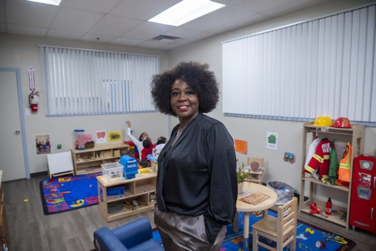 The operator of childcare center, a young black woman, is seen in the play room with desks and play areas behind her. 