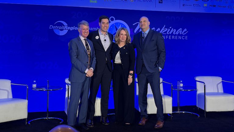 Three men and one women in business suits stand on a stage with a blue background with the words: Mackinac Policy Conference.