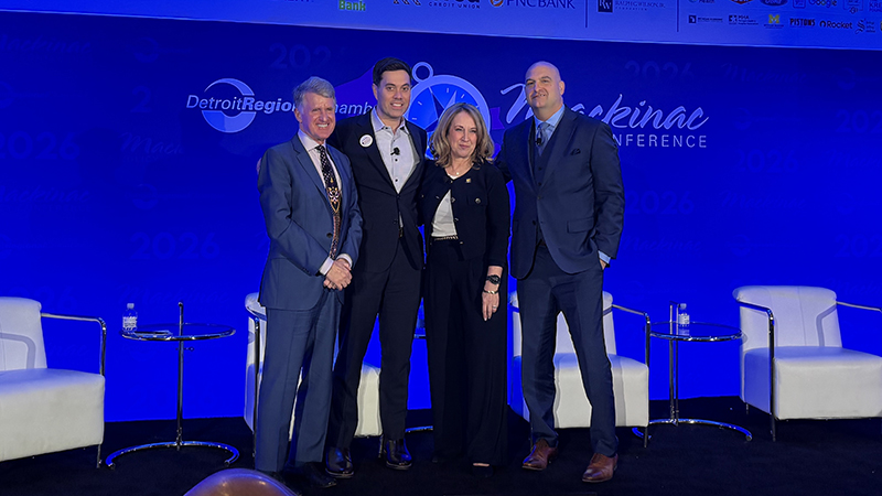 Three men and one women in business suits stand on a stage with a blue background with the words: Mackinac Policy Conference.