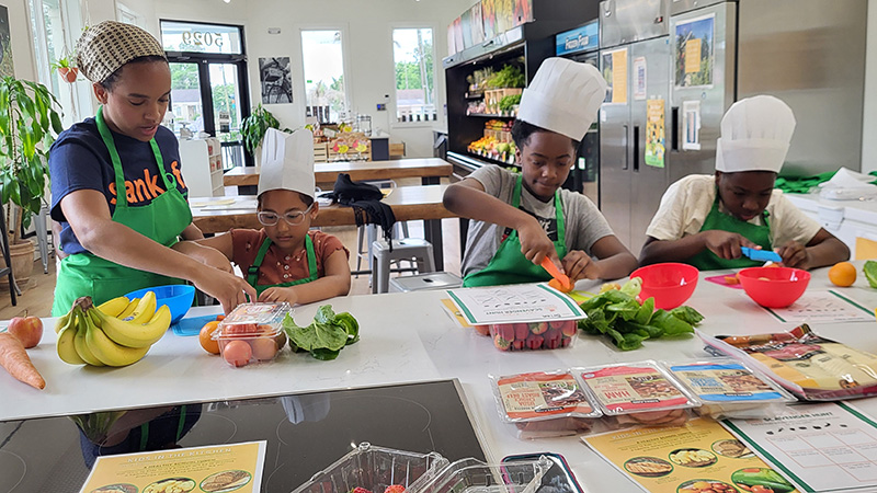 A woman in a Sankofa shirt and chef's hat is with three young kids also in chef hats as they are preparing meals 