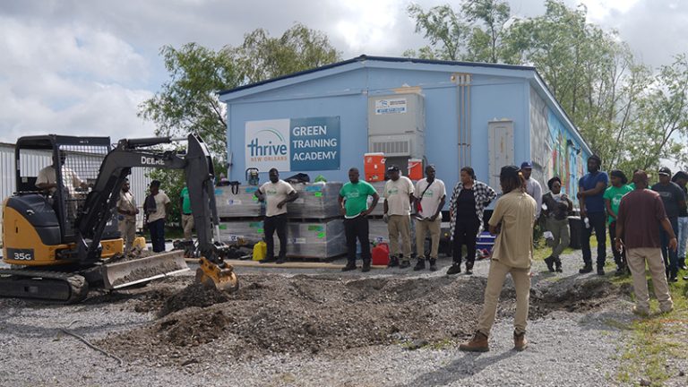 A group of people are gathered in front of a blue building with the sign Thrive New Orleans Green Training Academy. In front if them is a man in a excavator construction machine digging up the dirt.