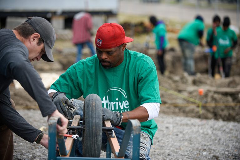 Two men (one wearing a green Thrive T-shirt and red hat and the other in a black hat and black T-shirt) working on repairing a wheelbarrow on an outside work project with a group of other workers in the background.